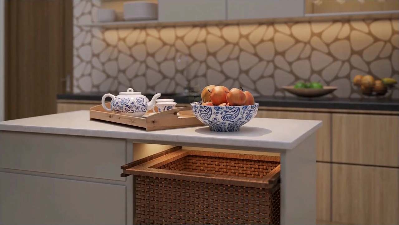 Close-up of a white quartz island with a paisley porcelain fruit bowl and tea tray, a pull-out rattan basket drawer glowing under LED strip light, and a softly blurred backdrop of lit pebble-mosaic backsplash and oak cabinetry.