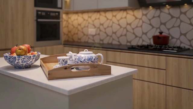 Shallow-depth-of-field kitchen detail: a blue paisley porcelain tea set on a pine serving tray and a bowl of red apples on a white quartz island, with warm LED light raking across an organic pebble-pattern backsplash, oak cabinets and a red cast-iron pot softly blurred behind.